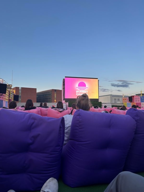 People seen from behind sitting in rows in front of a large cinema screen outdoors with city buildings and a blue evening sky in the distance