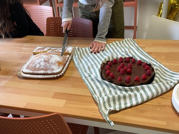A person's hands seen using a knife to cut into a lemon dessert on a table, while another uncut chocolate dessert with raspberries sits nearby