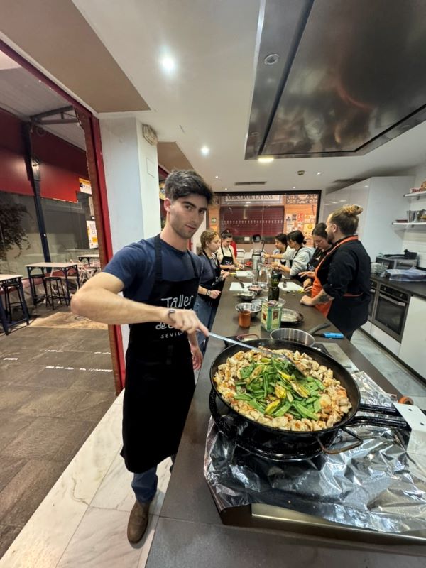 A study abroad student looking at the camera while stirring a large paella pan full of seafood and green vegetables