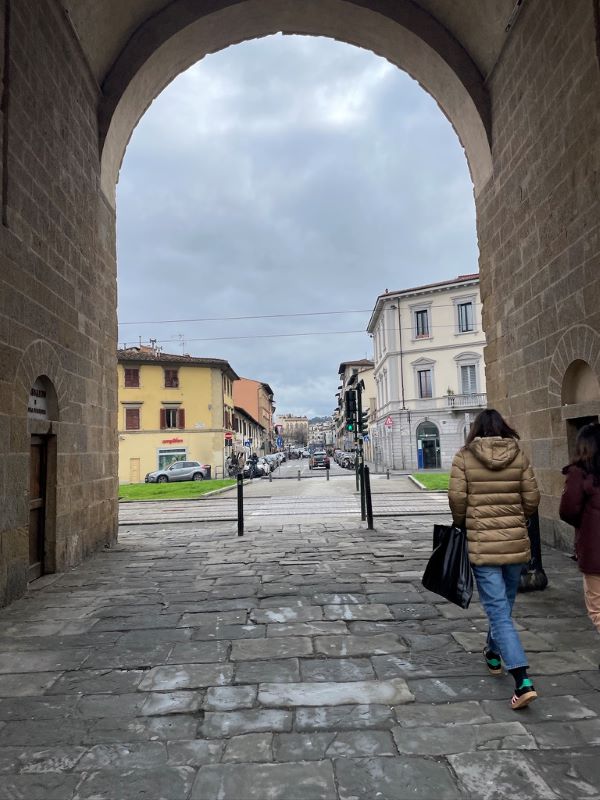 Two people seen from behind walking along a street underneath a stone archway with buildings in the distance