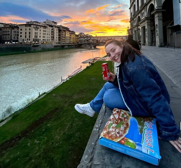 a study abroad student sitting along a river with pizza and a soda