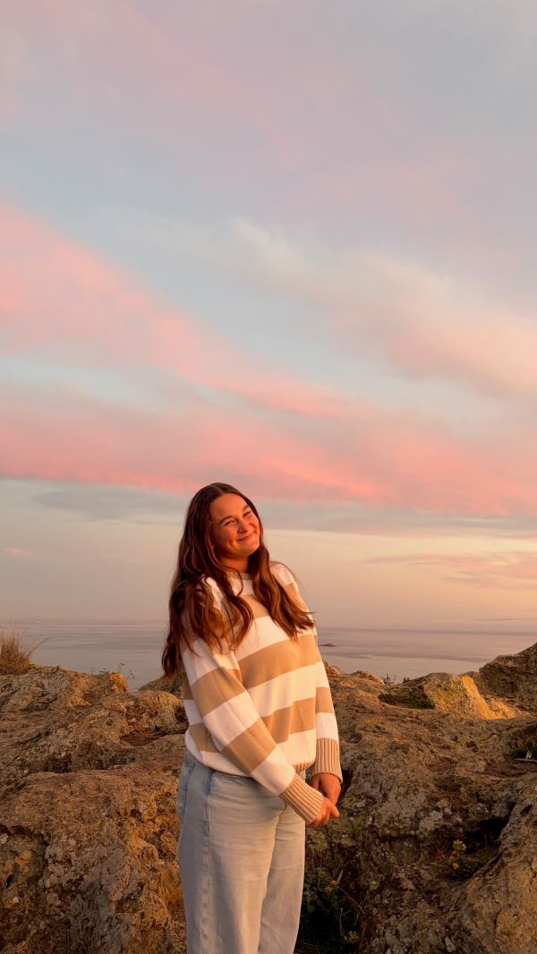 A study abroad student standing outdoors and holding their hands together in front of them while smiling at the camera