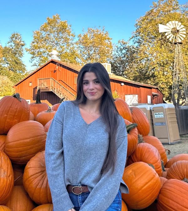 A study abroad student wearing a gray sweater and smiling at the camera outdoors in front of a large pile of pumpkins
