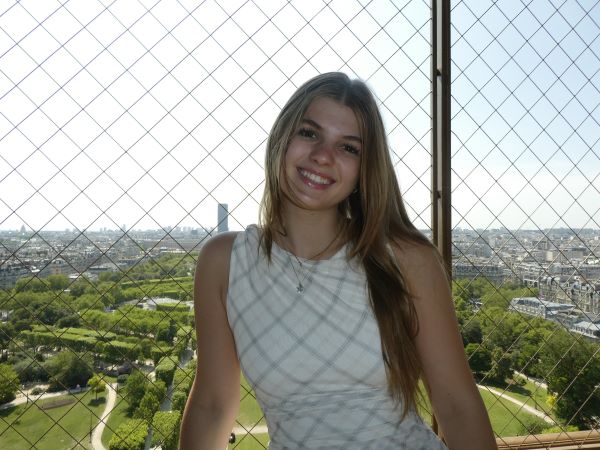 A study abroad student smiling at the camera in front of a fenced-in area high above a green space and city landscape in the background