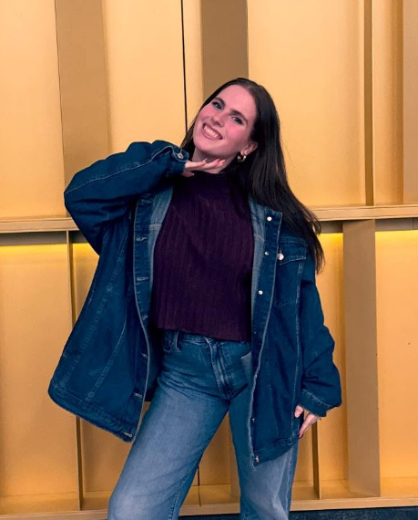 A study abroad student wearing jeans and a large jean jacket smiling at the camera with one hand held up underneath their chin