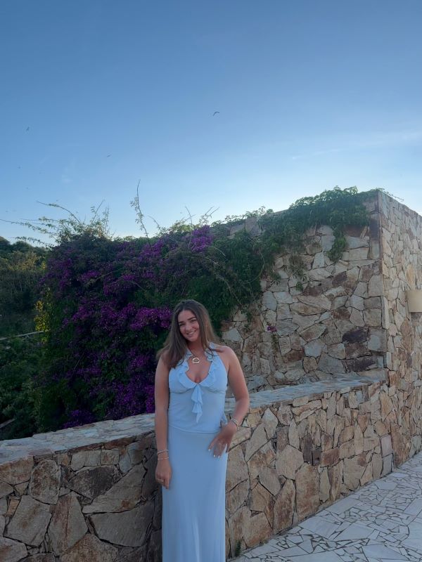A study abroad student wearing a blue dress smiling at the camera while standing outdoors in front of a stone wall