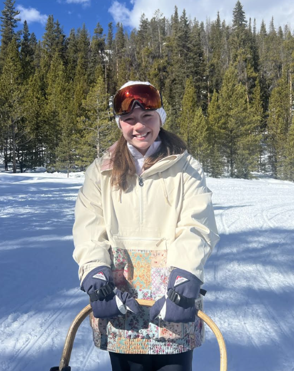 A study abroad student wearing a winter coat, hat, and gloves and holding on to the top of a sled while smiling at the camera in a snowy landscape