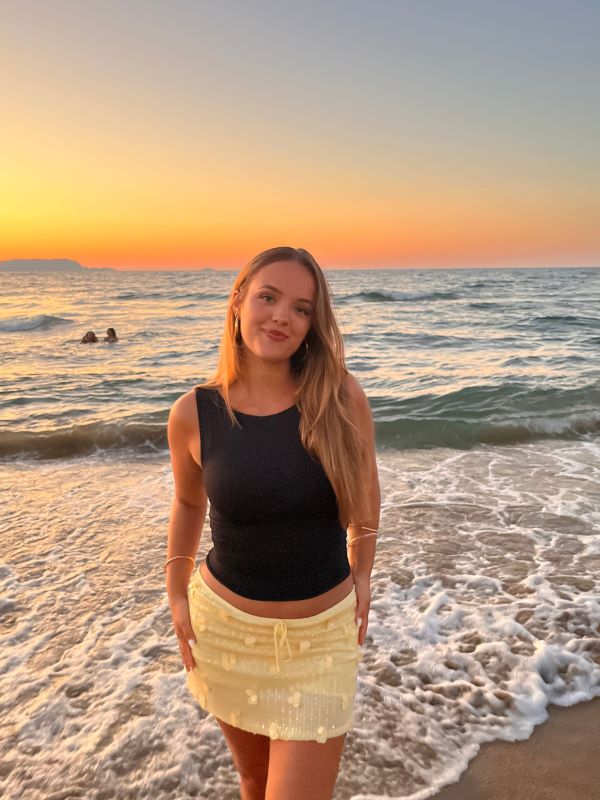 A study abroad student smiling at the camera while standing in front of an ocean at sunset