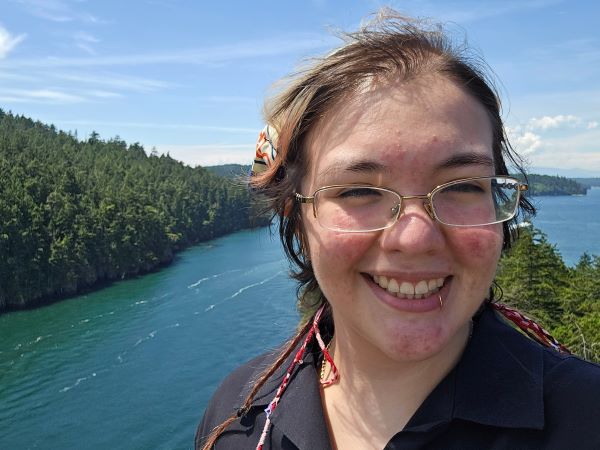 A study abroad student wearing glasses smiling at the camera in front of a large body of water hemmed in by a forest of trees