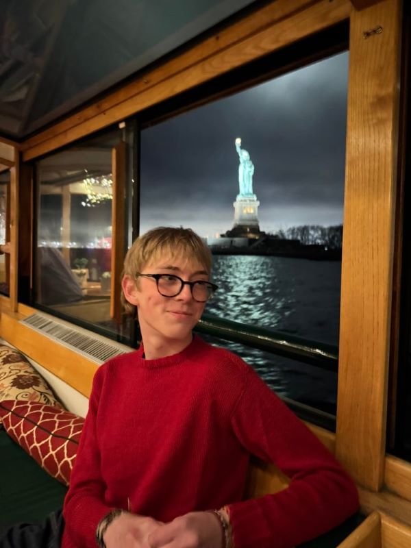A study abroad student sitting indoors and smiling while looking away from the camera with the Statue of Liberty visible in the window behind them